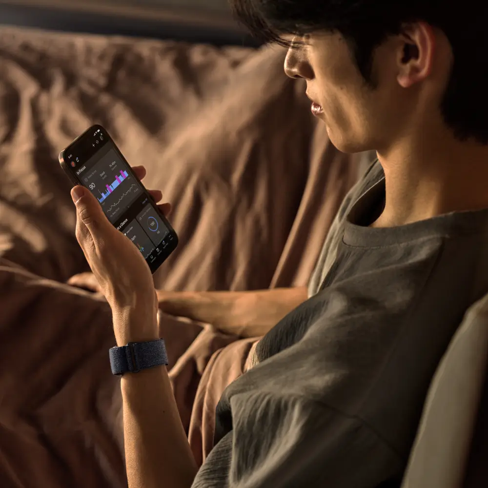 Ein Mann sitzt im Bett, trägt das Garmin Cirqa Smart Band in Kapitänsblau und prüft seine Fitnessdaten in der Garmin Connect Smartphone-App
