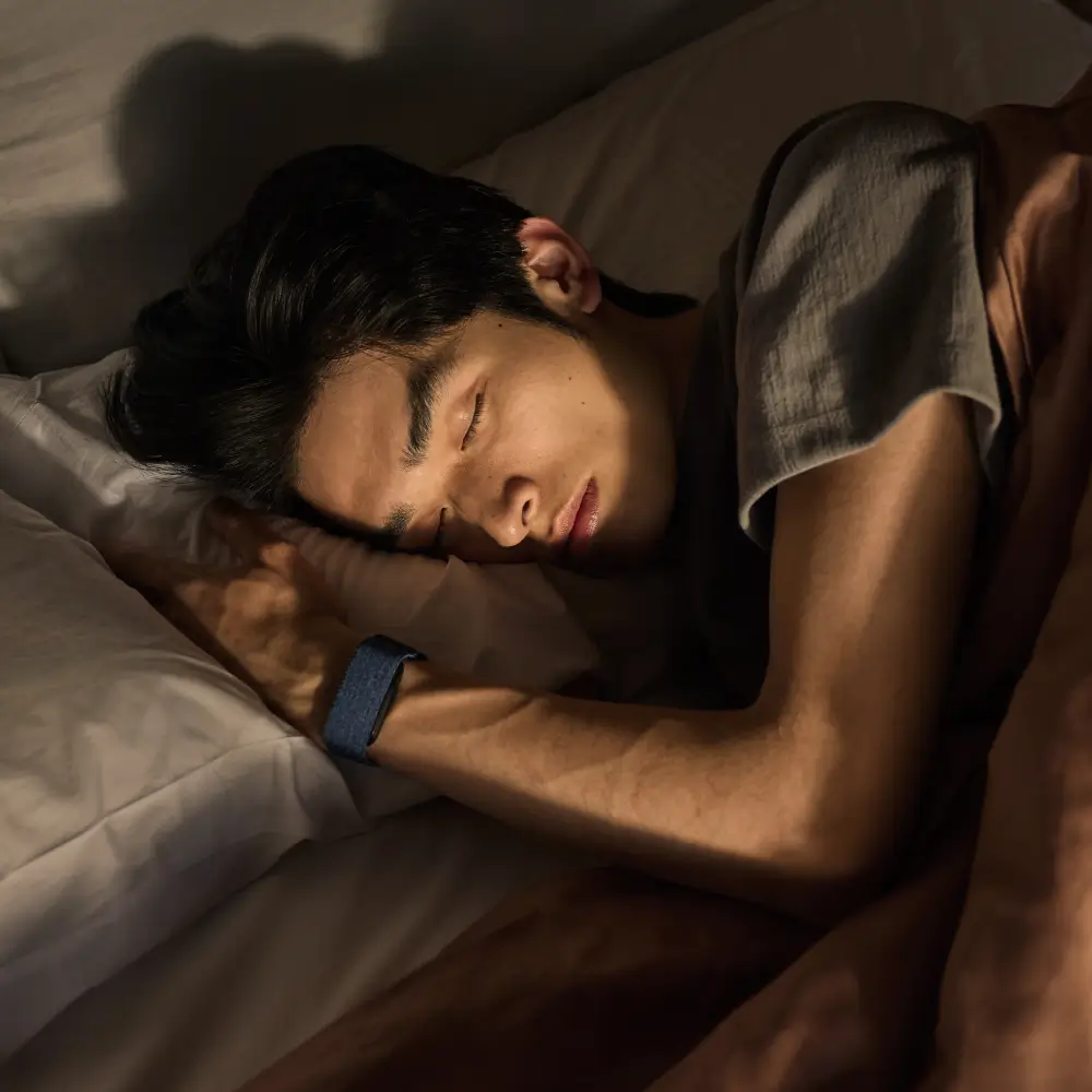 Ein Mann schläft im Bett und trägt das displaylose Garmin Cirqa Smart Band in Kapitänsblau zur automatischen Schlafanalyse am Handgelenk