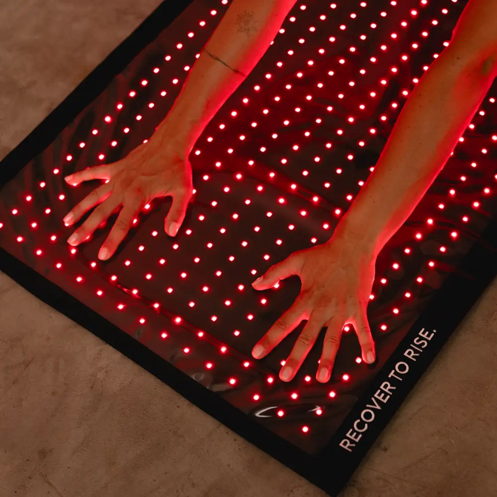 Nahaufnahme von zwei Händen auf einer leuchtenden Reboots Relight Mat Rotlichttherapie-Matte mit dem Schriftzug RECOVER TO RISE.