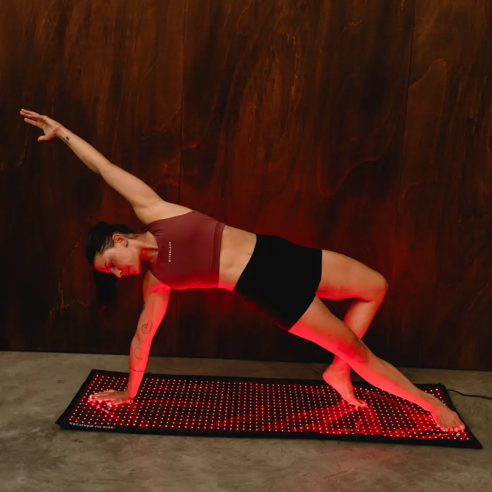 Eine Sportlerin hält eine Yoga-Position in der Seitenstütze auf der vollflächig rot strahlenden Reboots Relight Mat.