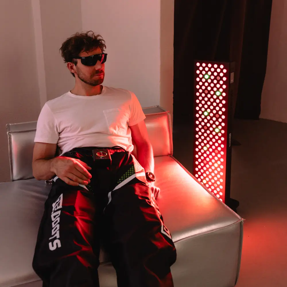 Ein Mann mit Laserschutzbrille nutzt die Reboots Regenerations-Boots im Sitzen, während im Hintergrund das Reboots Relight Pro Panel leuchtet.