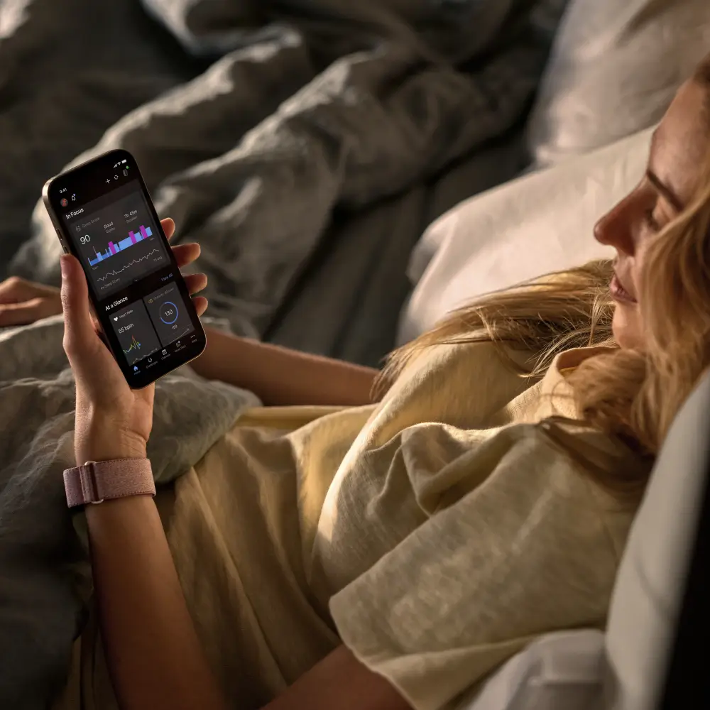 Eine Frau liegt im Bett, trägt das Garmin Cirqa Smart Band in Malve und prüft ihre Schlafanalyse in der Garmin Connect Smartphone-App