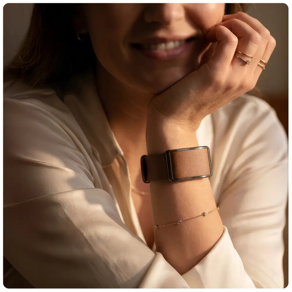 Lifestyle-Aufnahme einer lächelnden Frau, die ihr Kinn auf die Hand stützt. Sie trägt den POLAR Loop Fitness Tracker mit einem braunen Lederarmband, das als elegantes Accessoire perfekt mit feinem Goldschmuck und einer weißen Bluse harmoniert.